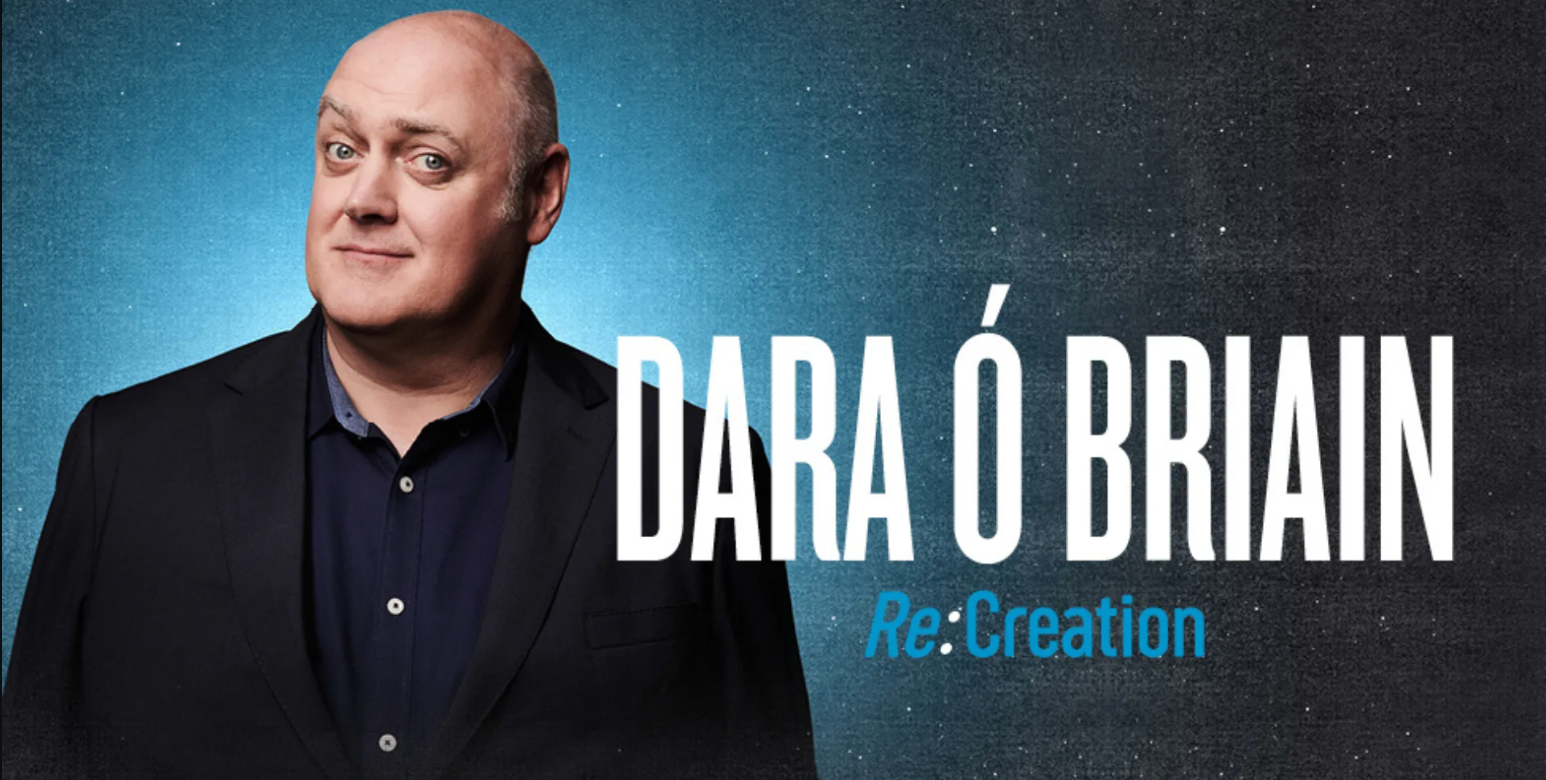 Dara O Briain stands with a quizzical expression on a pale blue background.