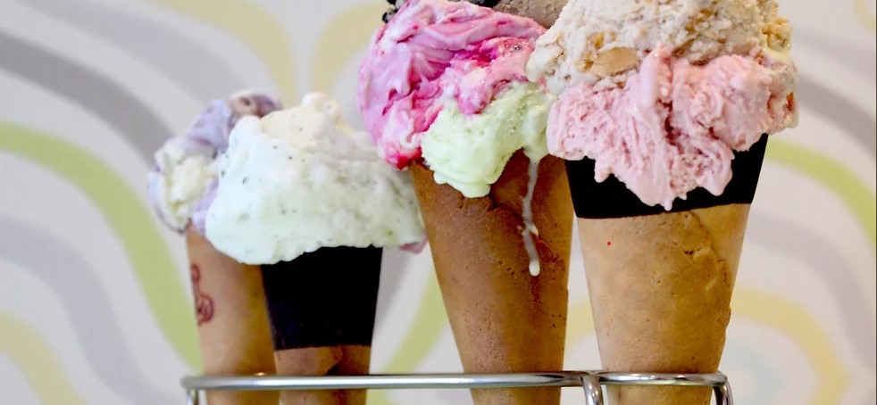 Four ice cream cones, each with a variety of flavors and toppings, are held in a metal stand. The ice cream flavors include a mix of colors such as white, pink, green, and purple, with some melting and dripping over the sides of the cones.
