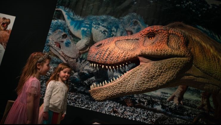 Two young children stand in awe, smiling at a large, realistic dinosaur exhibit featuring a detailed animatronic T-Rex with an open mouth. Colorful dinosaur illustrations adorn the wall behind them, adding a vibrant prehistoric atmosphere to the scene.
