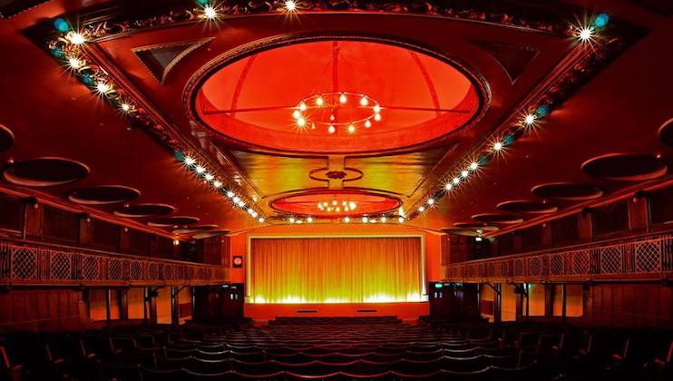 A beautifully lit theater interior with red and gold accents. The stage is illuminated with a drawn golden curtain. Rows of empty seats lead up to the stage, while the ceiling features intricate designs and circular light fixtures, adding elegance to the venue.