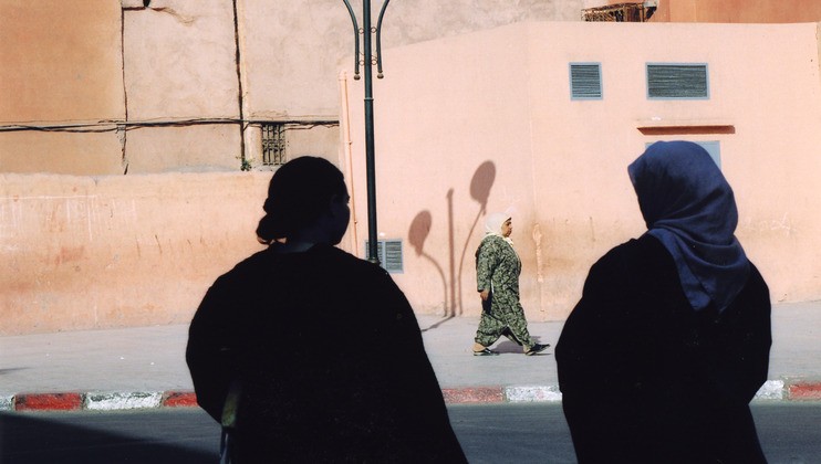 Silhouetted figures of two women are seen in the foreground, wearing dark clothing. In the background, a woman in green traditional attire walks past a peach-colored wall with a streetlamp casting shadows. The scene takes place on a sunny, urban street.