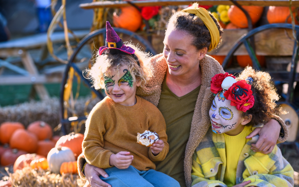 A mother smiles down at her two face-painted children eating Halloween themed cupcakes in front of a pile of pumpkins.
