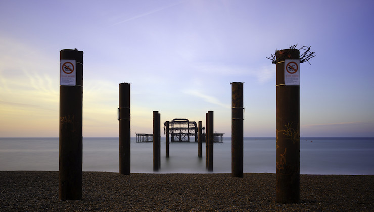 A serene image of a beach at twilight with the remains of a dilapidated pier stretching into the calm sea. Four rusted pillars with warning signs stand in the foreground, framing the skeletal structure of the pier against a backdrop of a soft, pastel-colored sky.
