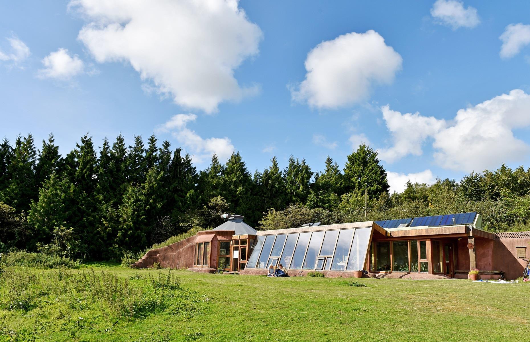 Stanmer Park's Earthship -  a sustainable eco lodge in stunning natural park, host of many spiritual and wellness retreats. A wooden house with slanted broad windows and solar panels before a lush forest.