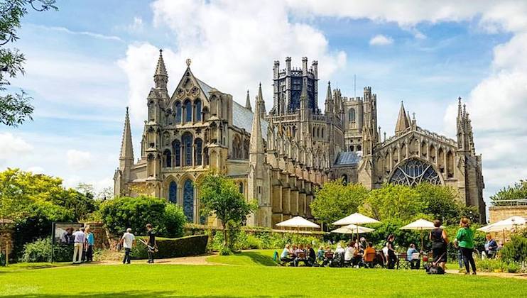 A majestic Gothic cathedral with pointed arches and intricate spires is set against a partly cloudy sky. In the foreground, people are enjoying the garden, some seated under white umbrellas on a lush green lawn. Trees and shrubs frame the scene.