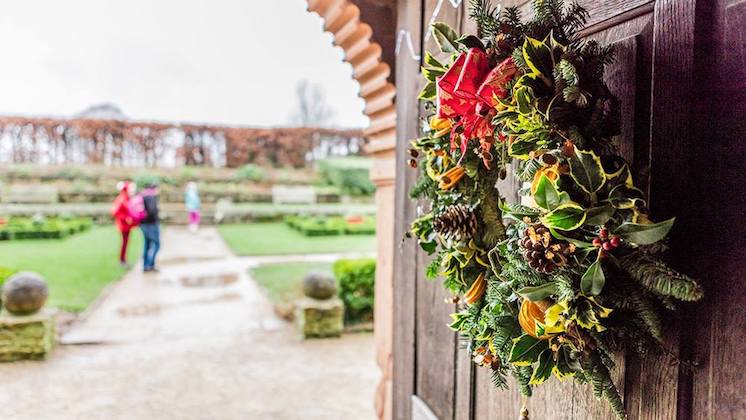 A decorated Christmas wreath with red ribbons, pinecones, and greenery hangs on a wooden door, leading to a garden path. In the background, a person in a red coat and a child walk along the wet pathway through the manicured garden.