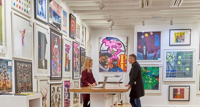 Two people are standing at a counter in an art gallery. The walls are covered with framed artwork of various styles and sizes. The gallery has a modern design with white walls and wooden beams on the ceiling. A laptop is open on the counter.