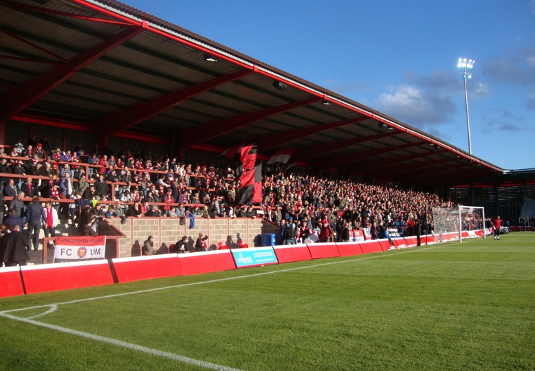A large crowd of football fans seated in a stadium stand, many waving red and black flags. The field is green with the corner and goal area visible. The stadium has a red roof structure and floodlights are lit against a clear blue sky.