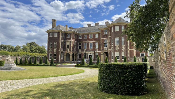 A large, historic brick mansion with tall chimneys and numerous windows standing in a well-maintained garden. The foreground features a circular driveway, lush green lawn, manicured hedges, and stone statues. The sky is partly cloudy with blue patches.