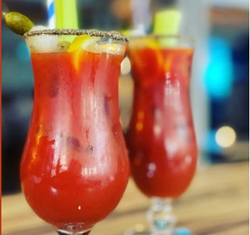 Close-up of two Bloody Mary cocktails in tall glasses on a wooden surface. Each glass has a salted rim and is garnished with a celery stalk and a lemon wedge. The drinks have a rich red color and appear refreshing, with a blurred background.
