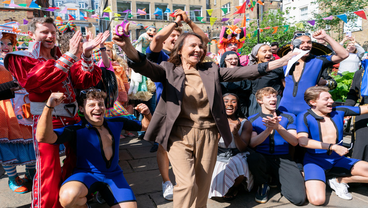 A jubilant crowd celebrates outdoors, featuring a woman in brown clothing dancing in the center. Surrounding her are people in colorful costumes and blue athletic outfits, with raised arms and joyful expressions. Bright bunting and a festive atmosphere are in the background.