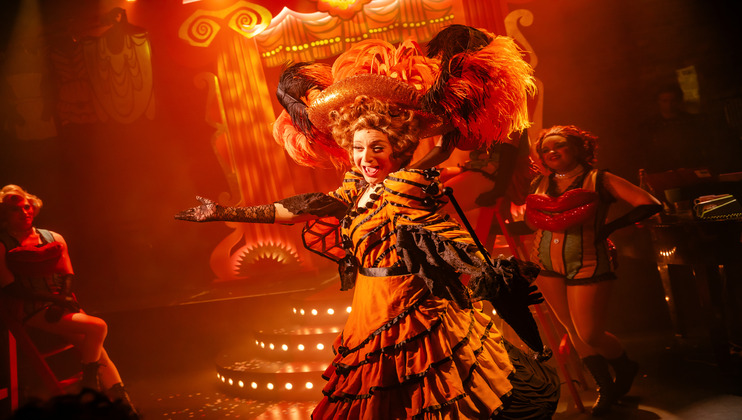 A performer in an elaborate, orange and black costume poses dramatically on stage, with a large, feathery headdress. The stage has bright, colorful lighting and a whimsical, carnival-like set in the background. Other performers in vivid costumes are visible.