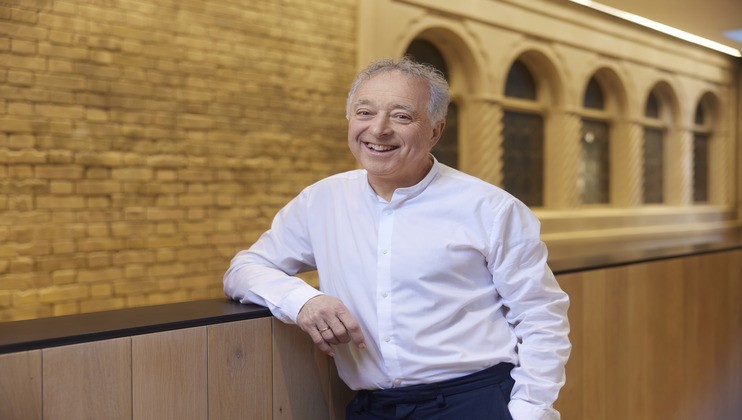 A smiling elderly man with white hair is leaning casually against a wooden railing with one hand. He is wearing a white button-down shirt. The background features a decorative indoor setting with brick walls and arched windows.