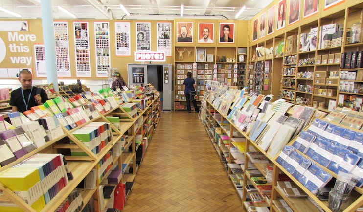 A well-lit bookstore with wooden shelves filled with books, magazines, and various other items. Two people browse through the materials. The walls display framed photos and large posters. A sign reading Photos hangs from the ceiling at the far end.