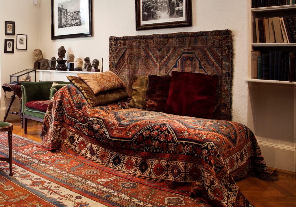 A room with a vintage chaise longue covered in an ornate, colorful patterned rug and adorned with assorted cushions. The floor features similar patterned rugs, and the walls are decorated with framed pictures. A bookshelf and bust sculptures are in the background.