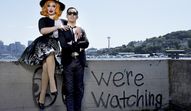 A person wearing a black hat, a wig, a black floral dress, and high heels sits on a concrete wall. Another person in dark sunglasses and a suit stands beside them. The wall has we're watching graffiti, and a skyline with the Space Needle is visible in the background.