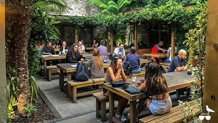 A lively outdoor garden seating area with people enjoying drinks and conversations at wooden picnic tables. The space features lush greenery, including vines, ferns, and trees, creating a cozy ambiance. The atmosphere is relaxed and social.