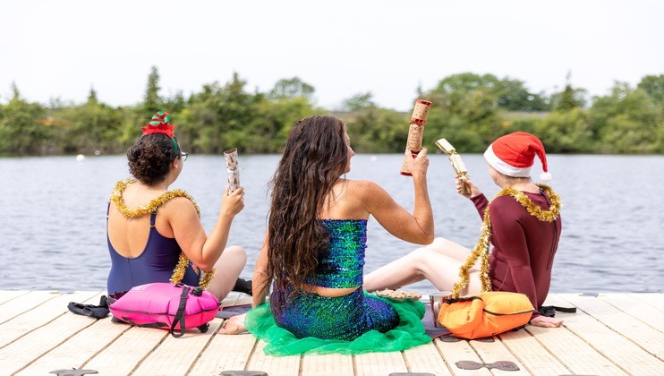 Three women in festive attire sit on a wooden dock by a lake. One wears a mermaid costume, another a Santa hat, and the third a reindeer headband. All have tinsel decorations and hold holiday crackers. The scene is cheerful with greenery in the background.