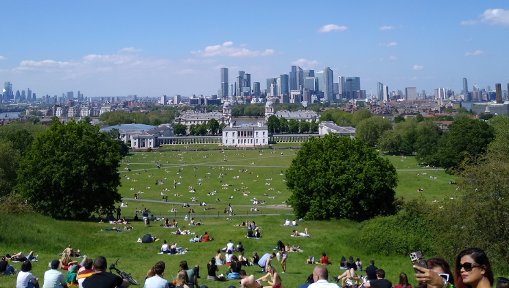 A wide shot of a park filled with people lounging and walking on a sunny day. In the background, a historical building with columns is visible, and beyond that, a modern city skyline with numerous skyscrapers. The scene is framed by lush green trees.