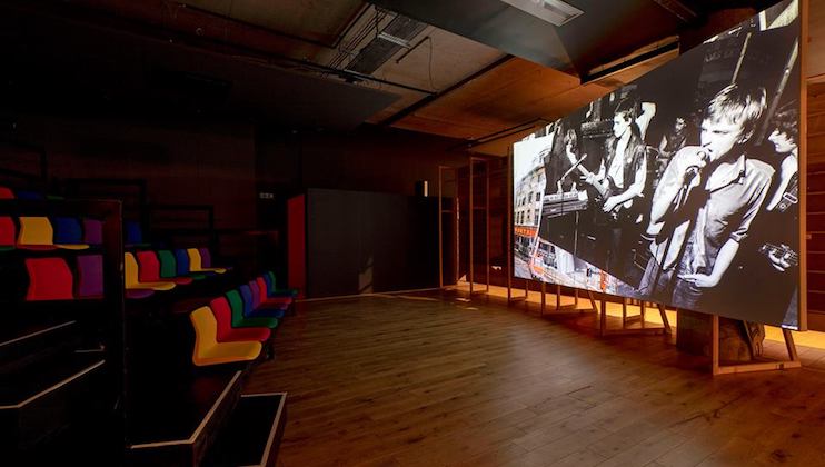 A dimly lit room features a large screen displaying a black-and-white image of a band playing instruments. The room has tiered seating with colorful, multi-colored cushions on the left and a wooden floor. The overall ambiance is modern and minimalist.