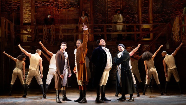 Cast of a musical performing on stage. Four performers stand prominently in front, with one pointing upward. Others are in the background with raised arms. The set includes wooden structures and ropes, suggesting a historical context.
