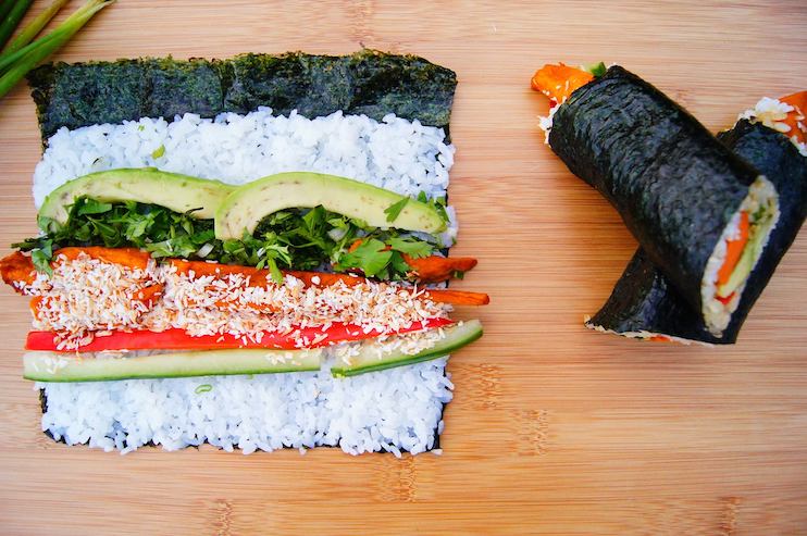 A sushi roll with nori, rice, avocado, cucumber, bell pepper, shredded carrots, and a sprinkle of sesame seeds is partially unrolled on a wooden board. Next to it is a cut sushi roll, revealing the vibrant fillings.