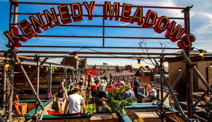 A lively rooftop bar named Kennedy Meadows with patrons enjoying food and drinks under clear skies. Neon sign reads LIBERTY in the background. The space features colorful seating and lush plants, creating a vibrant and relaxed atmosphere.