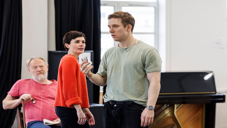 Three people are in a room: a seated older man with a white beard wearing a red shirt, a woman in an orange sweater looking towards the camera, and a younger man in a green shirt holding a smartphone, appearing to speak. A piano is in the background.