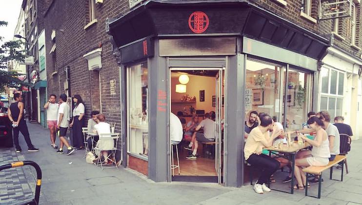 A bustling cafe sits at the corner of a brick building. People are seated both inside and outside at small tables, engaging in conversation. Some are standing on the pavement socializing. The entrance has large open windows, and the atmosphere is lively.