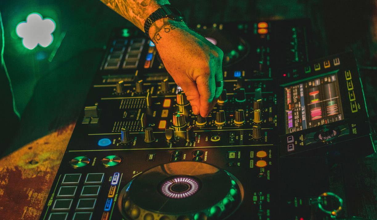 A DJ mixing tracks on a professional deck under green and yellow lights, creating a vibrant club atmosphere perfect for a Halloween house party.