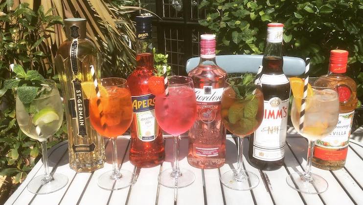 A selection of colorful cocktails in stemmed glasses displayed on a white table outdoors, each garnished with fruit and herbs. Behind the glasses are bottles of liqueurs, including St-Germain, Aperol, Gordon's Pink Gin, Pimm's, and Malibu. Green bushes in the background.