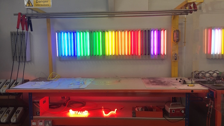 A workspace features a brightly lit display of colorful neon tubes arranged in a row on the wall, emitting a rainbow spectrum. Below, on a workbench, various tools, materials, and a glowing neon sign are visible, with caution signs indicating Danger for live busbars nearby.