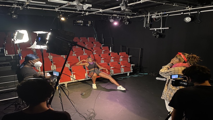 A person wearing a purple outfit poses dramatically on a red theater seat in an empty auditorium while three people with cameras and lighting equipment capture the scene. The person operating the key light is wearing a mask. The setting suggests a photo or video shoot.