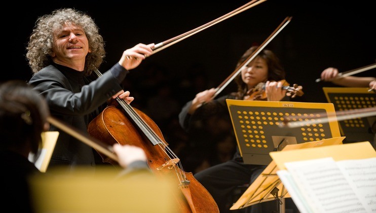 A cellist with curly hair passionately plays his instrument amidst an ensemble of violinists. Music stands with yellow sheets are visible, and the musicians appear focused on their performance in a dimly lit setting.