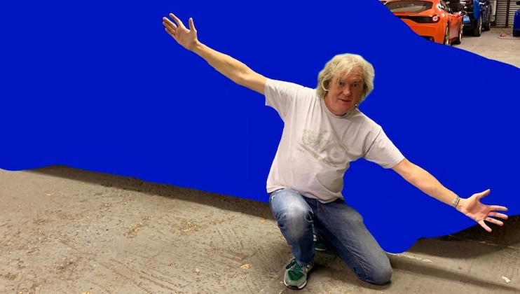 A middle-aged man with light-colored hair is kneeling on one knee and spreading his arms wide, posing in front of a blue object. He is wearing a light gray T-shirt, blue jeans, and green shoes. The background shows a garage with another car visible.
