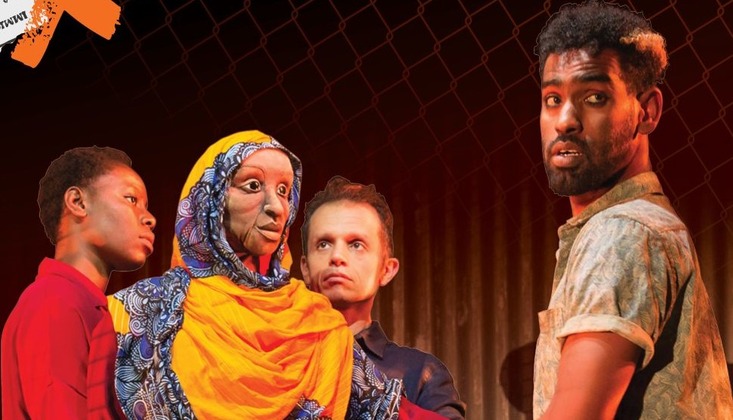 A diverse group of four individuals stands in front of a dark background with a chain-link fence. One person wears a patterned yellow and blue outfit with a headscarf, another wears a red shirt, the third wears a dark blue outfit, and the fourth wears a light shirt.