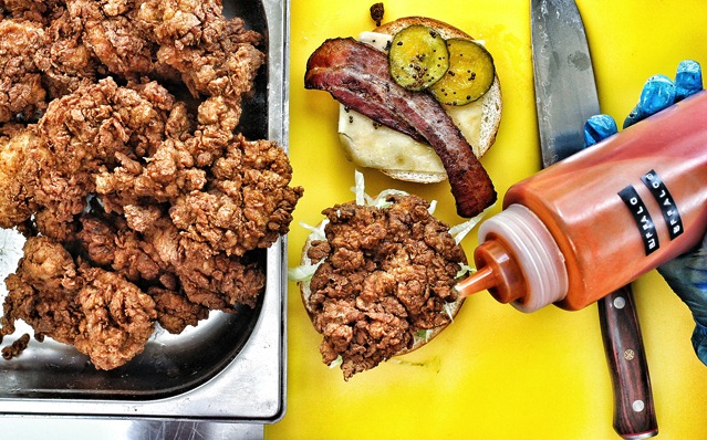 A tray of fried chicken pieces is on the left. Next to it, a sandwich is being assembled on a yellow surface with a bun, cheese, pickles, and a piece of bacon. Someone wearing a blue glove is squeezing sauce from a bottle onto the sandwich. A knife is nearby.