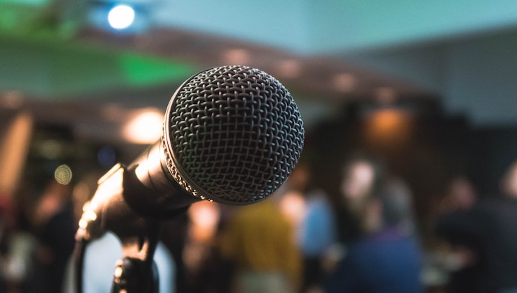 Close-up of a microphone on a stand, with a blurred background of people mingling in a dimly lit room. The microphone is prominently in focus, suggesting an event, speech, or performance is about to take place.