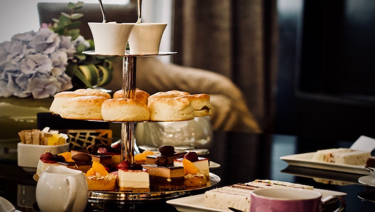 A two-tiered dessert stand holds scones and assorted pastries atop a table set for tea. There are teacups, a teapot, and various cakes. In the background, there are a vase with blue hydrangeas and blurred indoor decor, creating a cozy and elegant setting.