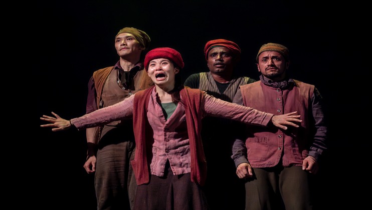 Four actors, dressed in rustic clothing and hats, stand on stage against a dark background. The actor in front, with an intense expression, extends both arms outward, while the three behind him display varied expressions from serious to contemplative.