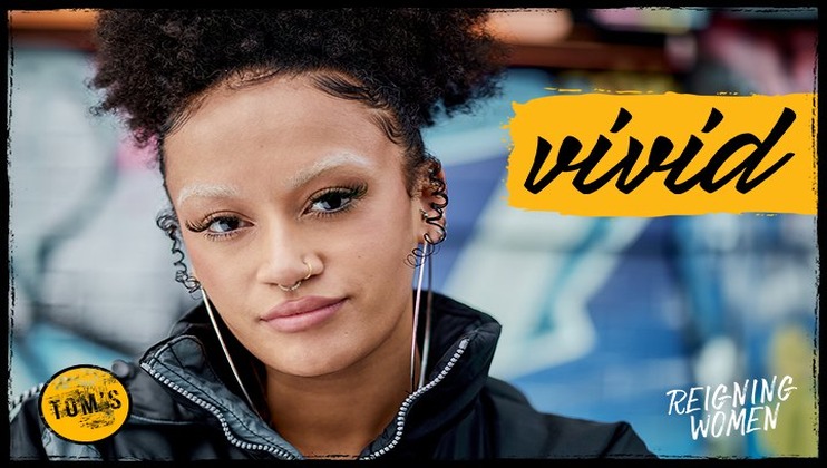 A person with curly hair and distinctive makeup looks at the camera against a graffiti-covered wall. They wear multiple ear piercings and a nose ring. The words vivid and Reigning Women appear on the image alongside a TOMS logo in the bottom left corner.