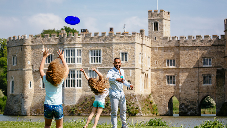 A man enjoys a sunny day outside a castle with two children. He throws a blue frisbee as the children leap with their arms outstretched to catch it. The castle, surrounded by a moat, sits in the background under a clear blue sky.