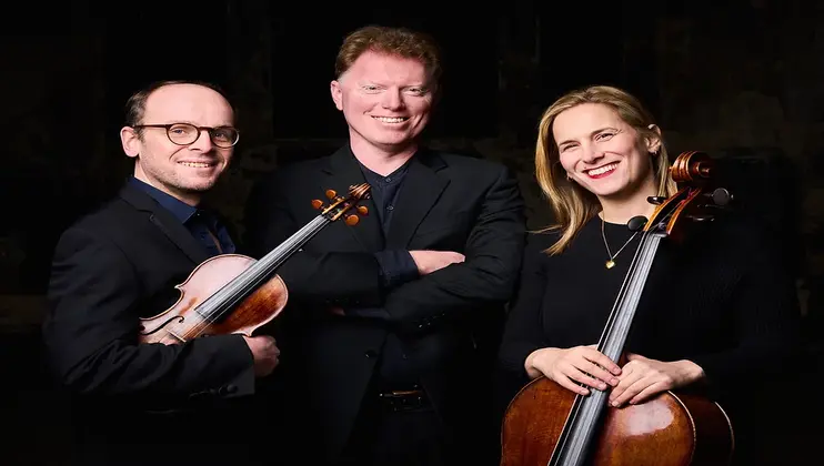 Three musicians, two men and one woman, pose together. The man on the left holds a violin, wearing glasses and smiling. The man in the center stands with arms crossed, also smiling. The woman on the right holds a cello and is smiling warmly. All wear dark clothing.