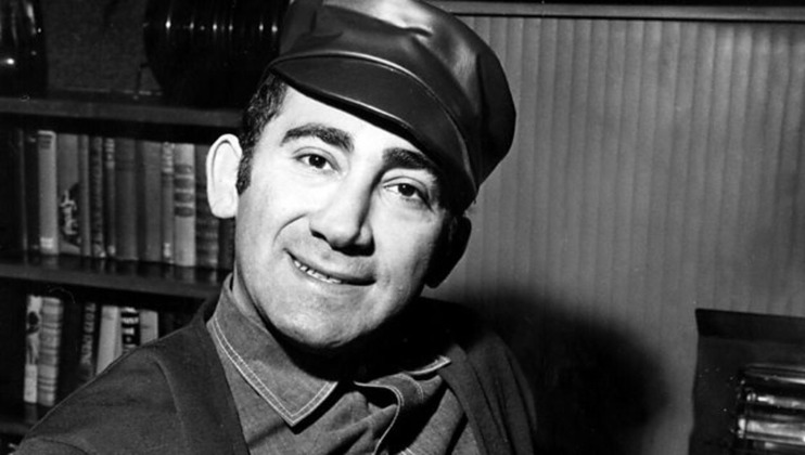 Black and white photo of a smiling man wearing a leather cap and a jacket. He is sitting in front of a bookshelf filled with books. The man is posed slightly at an angle, with a friendly expression on his face.