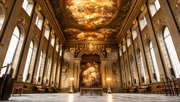 A grand hall with ornate golden architecture and numerous arched windows. The ceiling and walls are adorned with intricate fresco paintings. The hall features a marble floor and wooden benches along the sides, leading to a prominent, elaborately decorated central alcove.