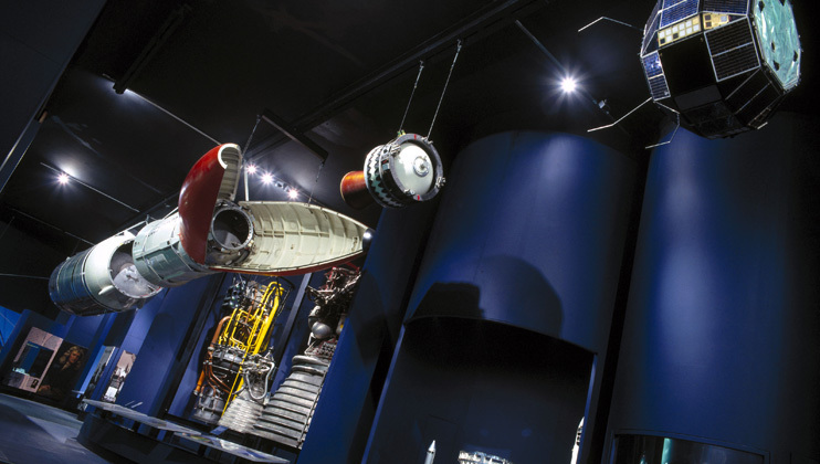 A museum display of various aerospace artifacts, including a spacecraft, satellite, and a rocket engine. The dark, dimly lit interior enhances the futuristic ambiance. The artifacts are suspended from the ceiling and showcased against a backdrop of vertical blue panels.