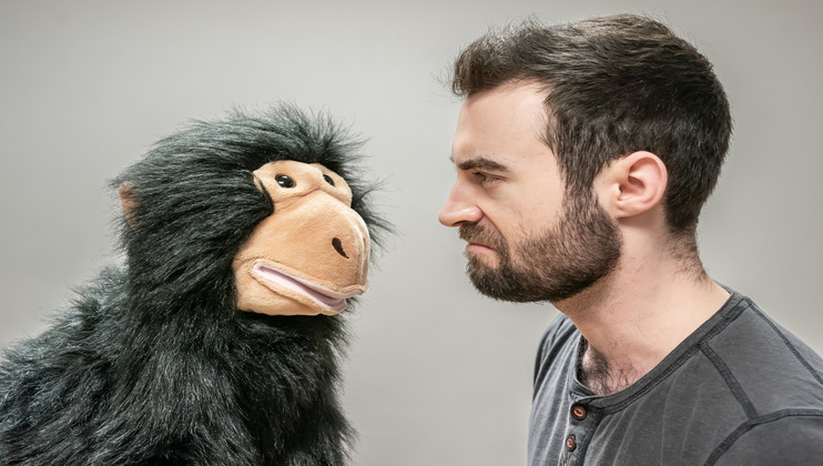 A bearded man with short dark hair glares at a stuffed black and tan monkey puppet. The puppet's face and the man's face are close to each other, with the man looking serious and the puppet's facial expression appearing neutral.