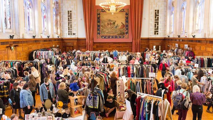 A bustling indoor market scene in a grand hall with high ceilings and ornate windows. Numerous stalls display a variety of vintage clothing and accessories. Shoppers are browsing and interacting with vendors, creating a lively and vibrant atmosphere.