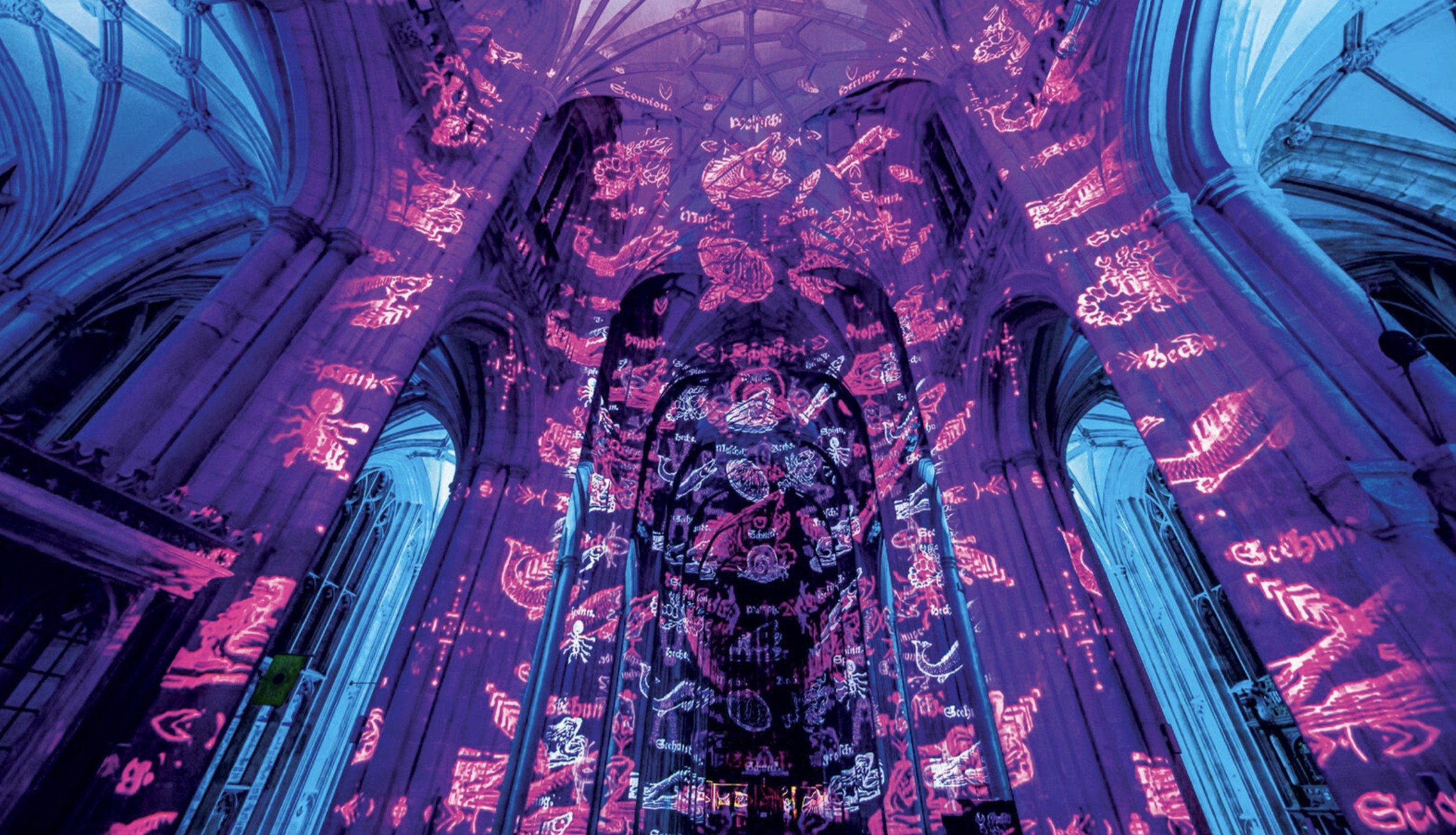A cathedral interior is illuminated with vibrant purple and blue light projections, featuring intricate designs and patterns on the arched ceiling and pillars, creating a mesmerizing and ethereal atmosphere.