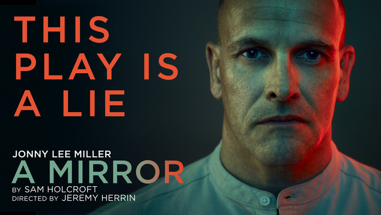 A serious-looking man with a shaved head is lit with dramatic red and green lighting. The text reads, This Play is a Lie. At the bottom, it says Jonny Lee Miller - A Mirror by Sam Holcroft, directed by Jeremy Herrin. The man wears a buttoned-up shirt.